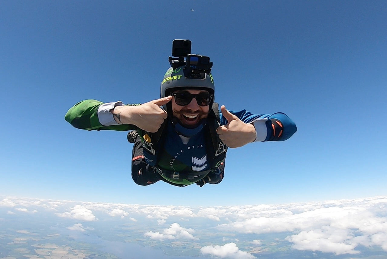 Licensed skydiver with mustache wearing a videography helmet giving two thumbs up while in freefall over Parachute Ottawa in Arnprior, ON