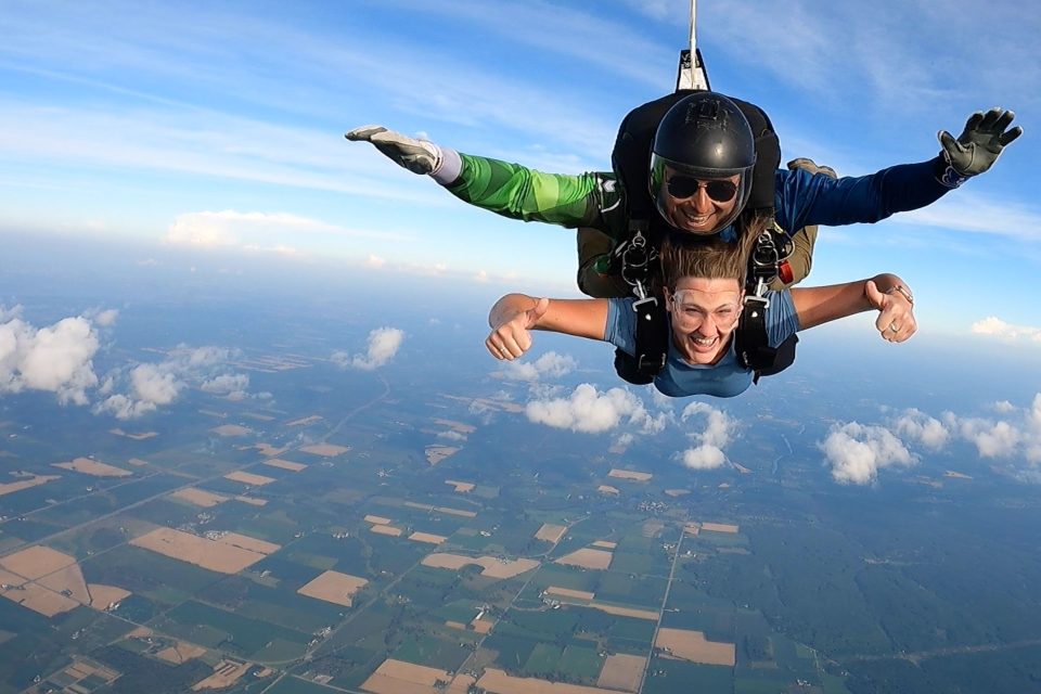 Female tandem skydiving student in blue shirt giving two thumbs up to camera while in freefall