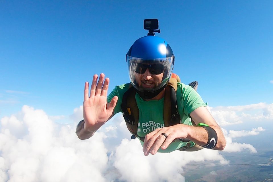 Experienced skydiver in freefall wearing a blue camera helmet and waving at the camera