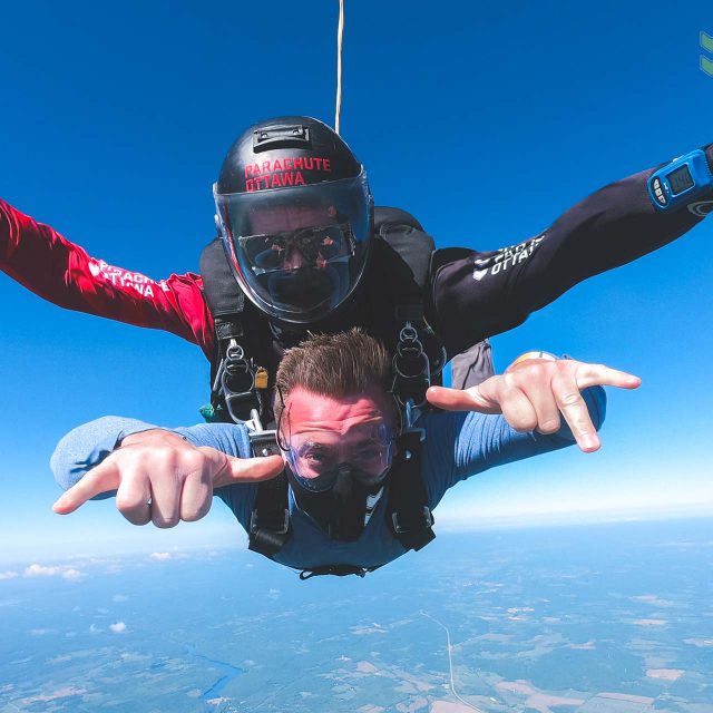 Male tandem skydiving student wearing a mask and instructor in freefall giving rock on signal with hands